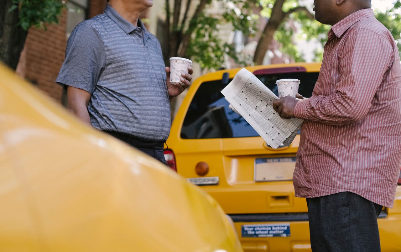 Crop anonymous multiracial taxi drivers with takeaway coffee and newspaper speaking near yellow cars in town